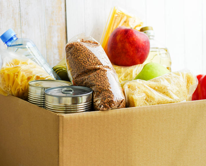 A cardboard box with food in, such as tins, a bottle of water, pasta, and fresh fruit and vegetables. 