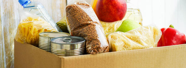 A cardboard box with food in, such as tins, a bottle of water, pasta, and fresh fruit and vegetables. 