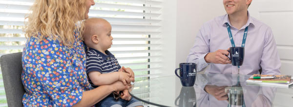 A male member of staff sat down at a dining room table drinking a cup of tea, talking to a resident and her young child. 