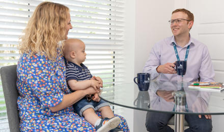 A male member of staff sat down at a dining room table drinking a cup of tea, talking to a resident and her young child. 