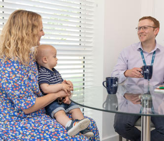 A male member of staff sat down at a dining room table drinking a cup of tea, talking to a resident and her young child. 