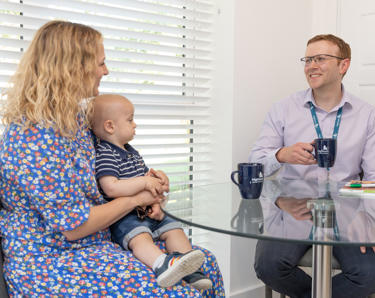 A male member of staff sat down at a dining room table drinking a cup of tea, talking to a resident and her young child. 