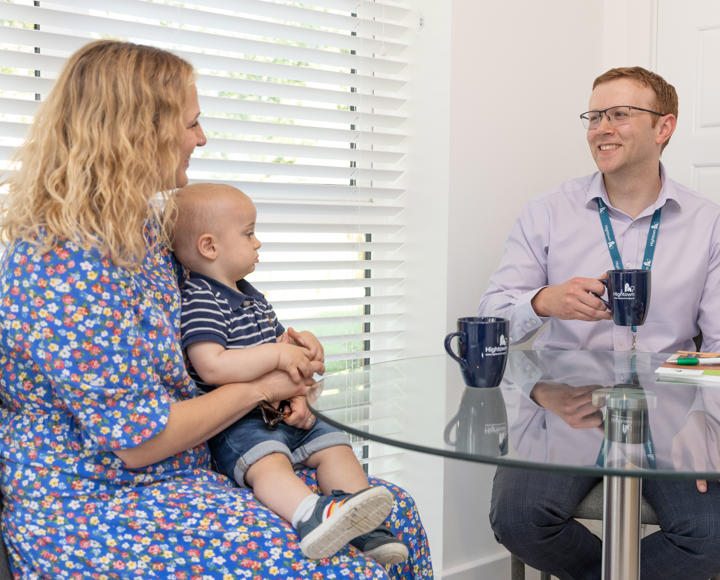 A male member of staff sat down at a dining room table drinking a cup of tea, talking to a resident and her young child. 
