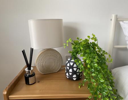An oak bedside table with a reed diffuser, lamp and false fern