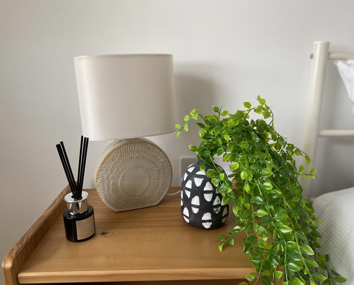 An oak bedside table with a reed diffuser, lamp and false fern