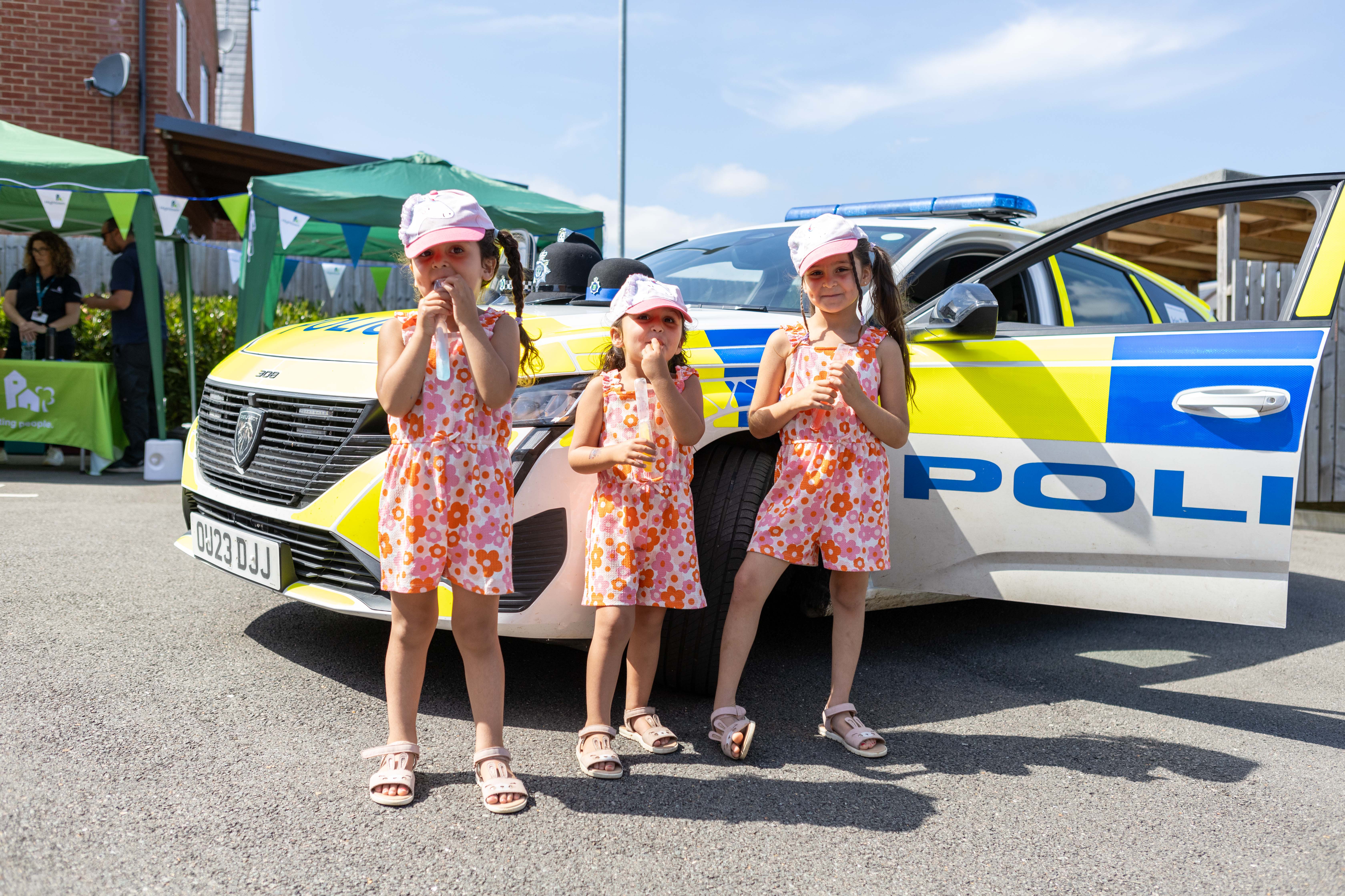 Three young children, stood in front of a police car on a sunny day, for a resident involvement open day.