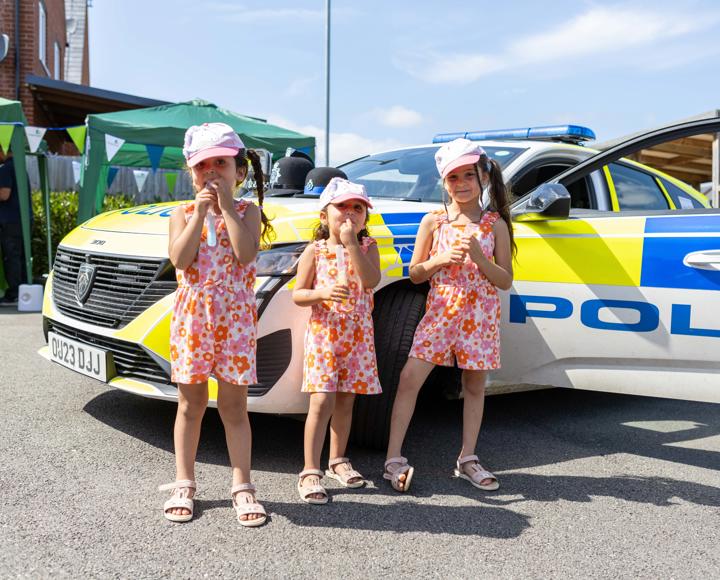 Three young children, stood in front of a police car on a sunny day, for a resident involvement open day.