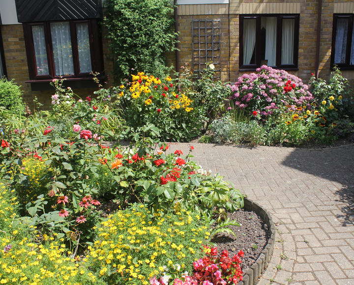 The front, ground floor of the outside of a block of flats. There is a large garden area with lots of colourful flowers planted in the ground.