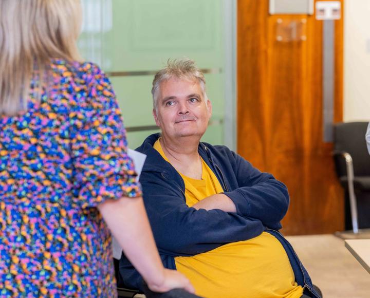 A man in a yellow tshirt and blue hoody, smiling, looking up at a woman in a colourful dress