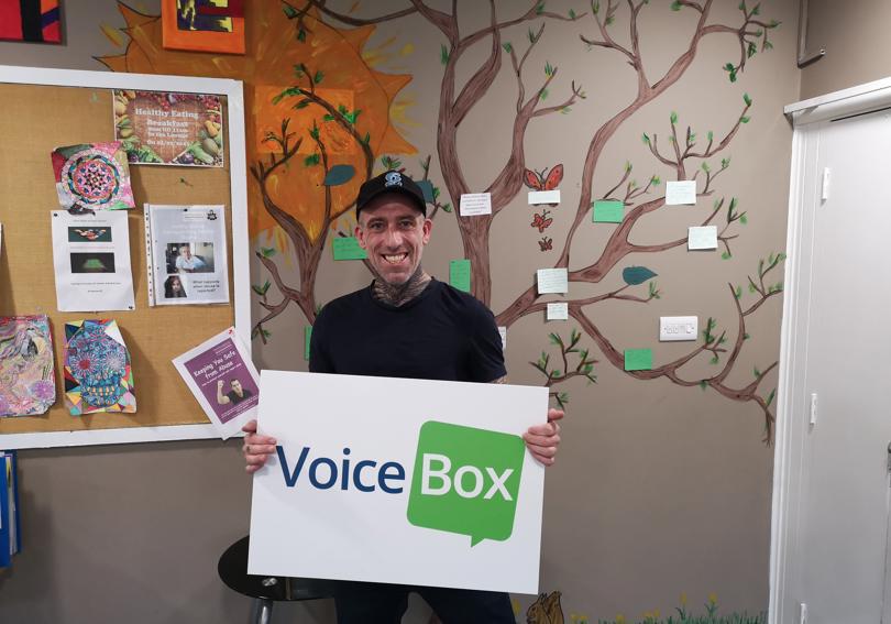 A man standing in front of a painting of a tree, holding a sign that says 'VoiceBox'
