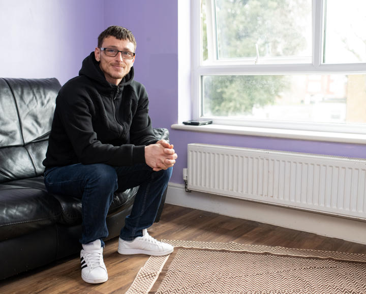 Hightown resident sitting on a sofa in his new home, with a black leather sofa and purple painted walls. 
