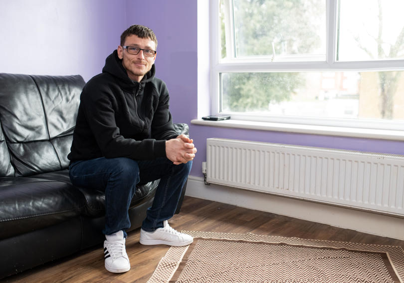 Hightown resident sitting on a sofa in his new home, with a black leather sofa and purple painted walls. 