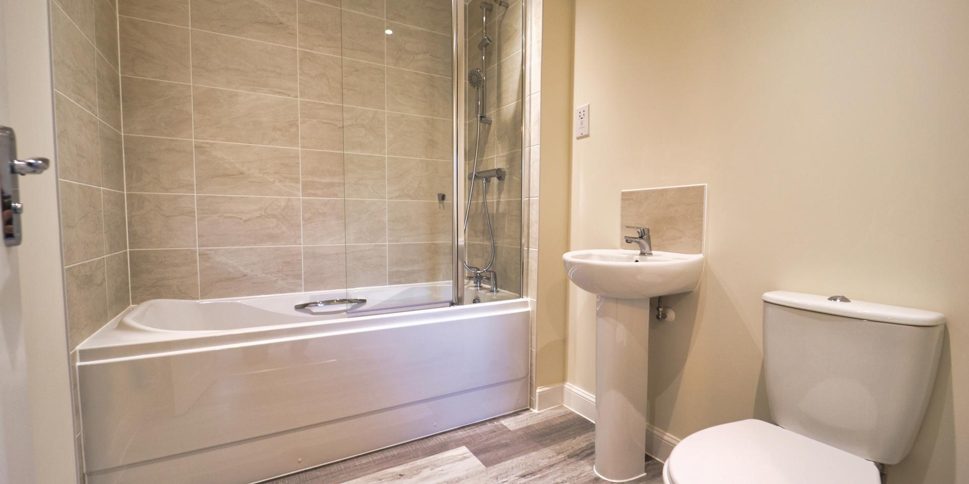 A white suite with shower over bath and shower head attached to the taps. There's a glass shower screen and pedestal sink and toilet