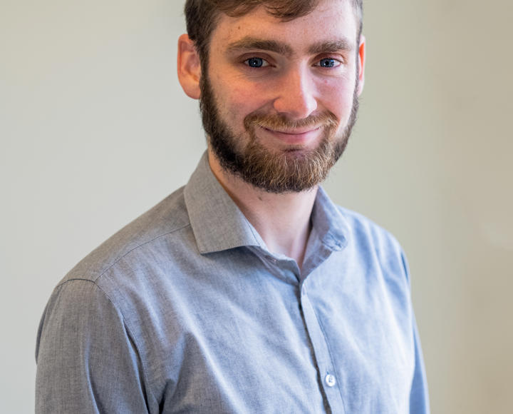 A headshot of a young male graduate trainee. He has brown hair and facial hair, with blue eyes. He is wearing a grey shirt.