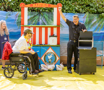Man in a wheelchair, female staff member and a male magician cheering with his hand in the air, in front of a puppet show